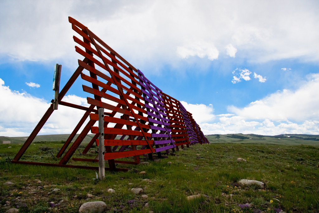 snow fence in a picturesque landscape with rocks and a meadow in teh foreground and clouds and epic mountains in the distance the sections of the snow fence are painted laternately red and purple